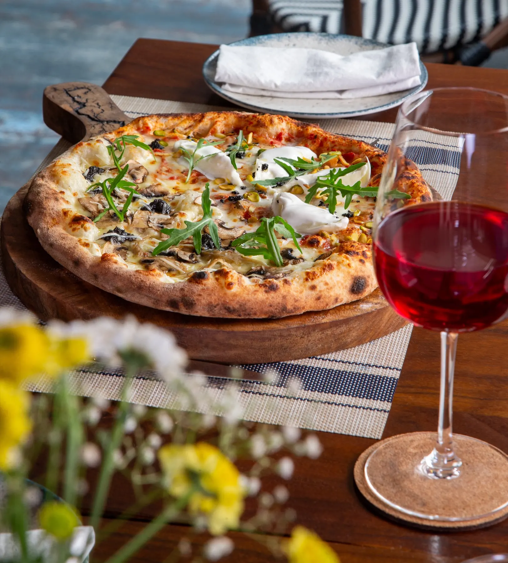 A wood-fired pizza with mushrooms, burrata and rocket beside a glass of red wine on a sunlit table at Rosmarino, Indiranagar.