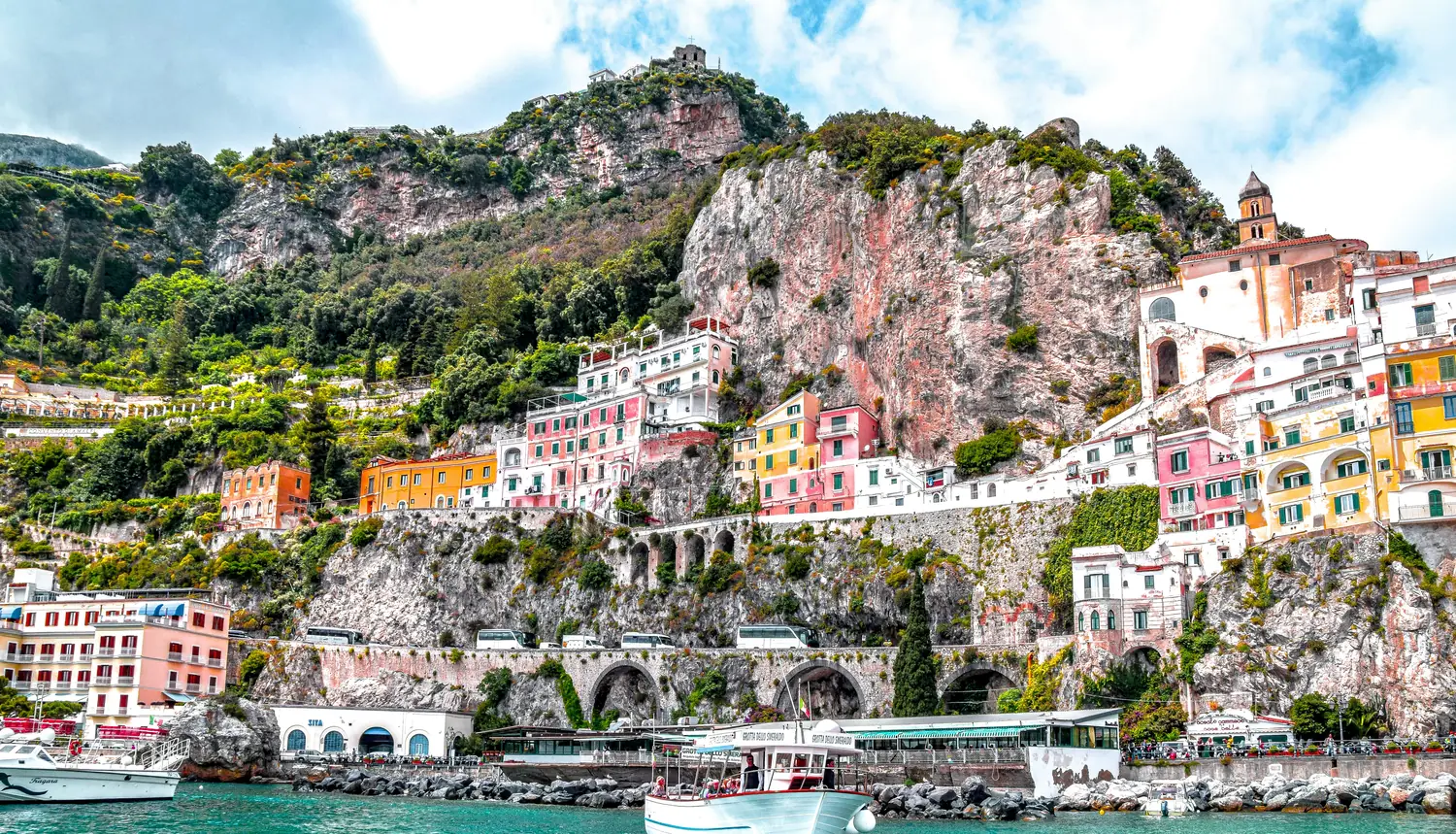 Amalfi town on a bright sunny day — colourful cliffside buildings above the turquoise Mediterranean, seen from the water