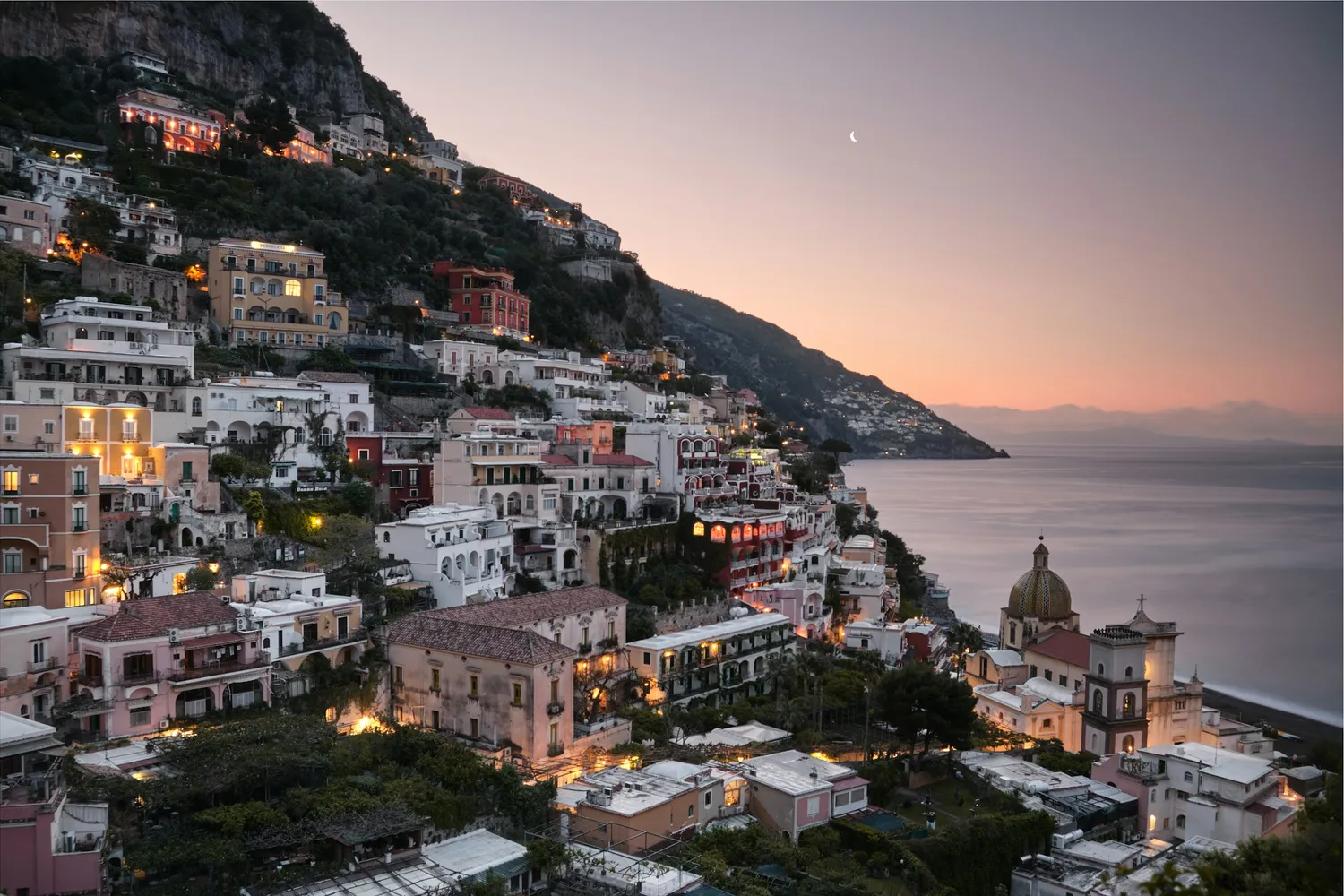 Positano on the Amalfi Coast at dusk — pastel cliffside houses glowing with warm light above the Mediterranean